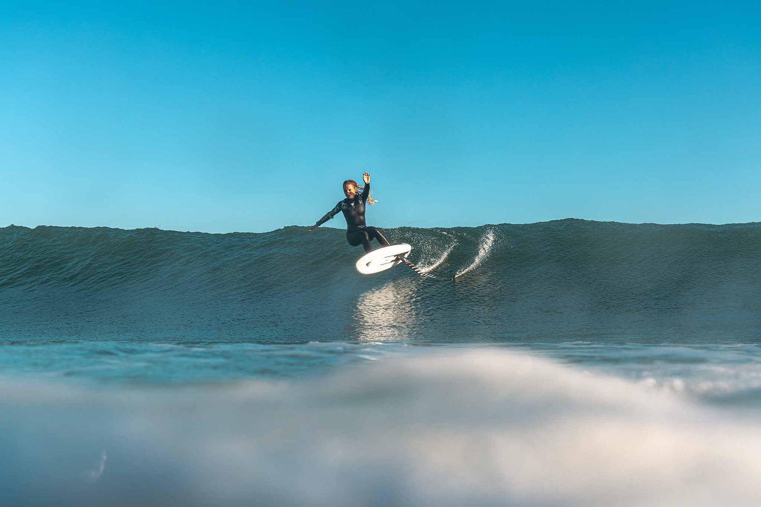 A female surfing on a wave with clear blue sky using the Armstrong Foils MA MkII Front Foil 