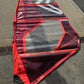A red and black windsurfer sail with a transparent center panel, laid out on a concrete surface outdoors. used Severne 2026
