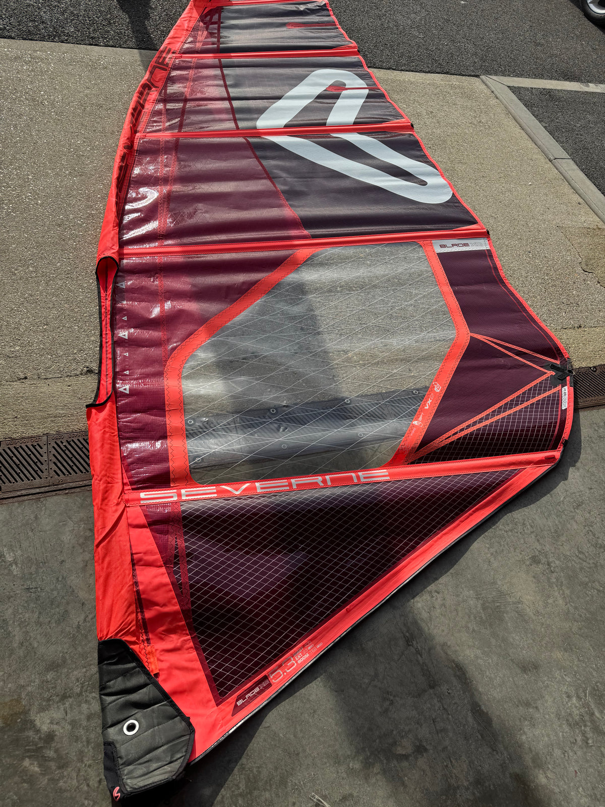 A red and black windsurfer sail with a transparent center panel, laid out on a concrete surface outdoors. used Severne 2026
