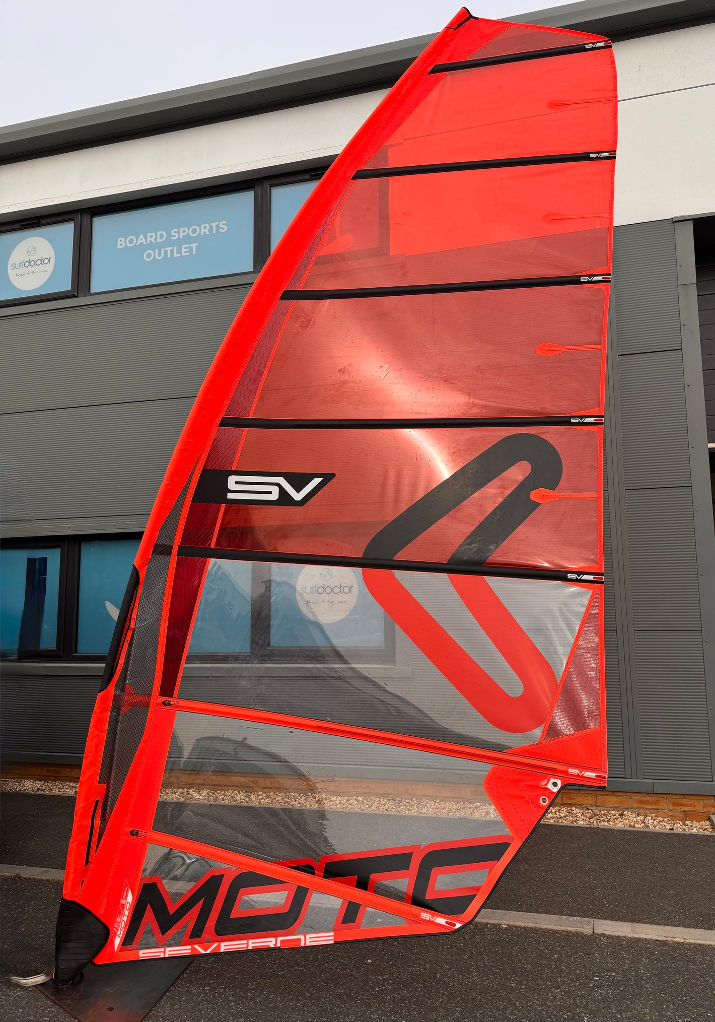 Red windsurf sail with 'SV' and 'MOTO' branding in front of a building.